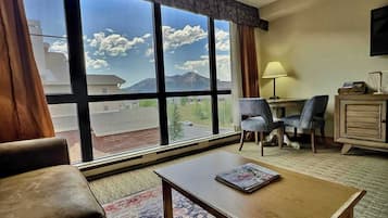 A hotel room with a large window offering a mountain view, a wooden coffee table, a sofa, a desk with a lamp, and a small cabinet.