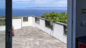 A balcony with a stone floor, a potted plant, and a view of the sea and distant mountains.