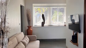 A spacious living area with a beige sofa, a large window with blinds, a television mounted on the wall, and a potted plant.