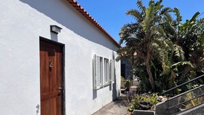 A white building with a brown door and a window with shutters. There is a concrete pathway leading to a set of stairs.