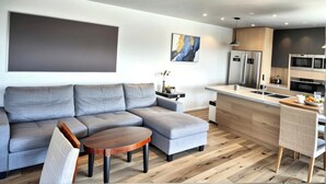 A modern living room with a grey sectional sofa, a wooden coffee table, and a kitchen area with a stainless steel refrigerator and microwave.