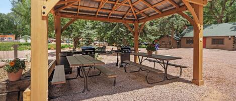 A covered picnic area with wooden benches and tables, surrounded by gravel and trees.