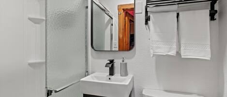 Un baño con mampara de vidrio, un lavamanos blanco con grifo negro, inodoro y perchero con toallas blancas.
