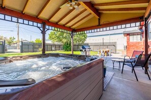 A hot tub under a covered patio with a wooden ceiling, outdoor furniture, and a barbecue grill in the background.
