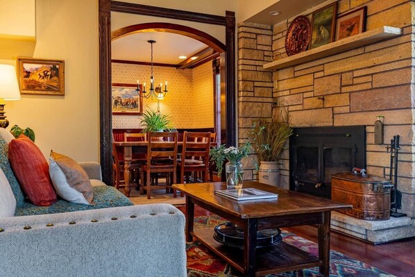 A cozy living room with a fireplace, a dining area with wooden chairs, and a sofa with cushions.