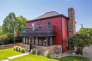 A two-story house with a stone chimney, a balcony with a railing, and a flagpole with an American flag.