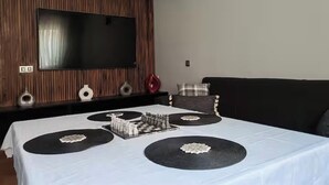 A modern living room with a large flat-screen TV mounted on a wooden wall, a black sofa, and a dining table set with chess pieces.