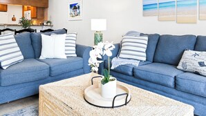 A modern living room with a blue sofa set, a coffee table with a plant, and artwork on the walls. - Coastal Comfort Steps from Cocoa Beach Pier W/ Heated Pool, BBQ Grills & Private Balcony (Cocoa Beach)