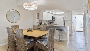 Een eethoek met rieten stoelen en een houten tafel, een keuken met witte kasten en een magnetron, en een grote spiegel aan de muur.