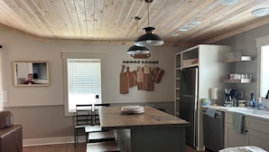 Une cuisine moderne avec un plafond en bois, un îlot central doté de tabourets, des ustensiles de cuisine suspendus, un réfrigérateur et une fenêtre avec des stores.