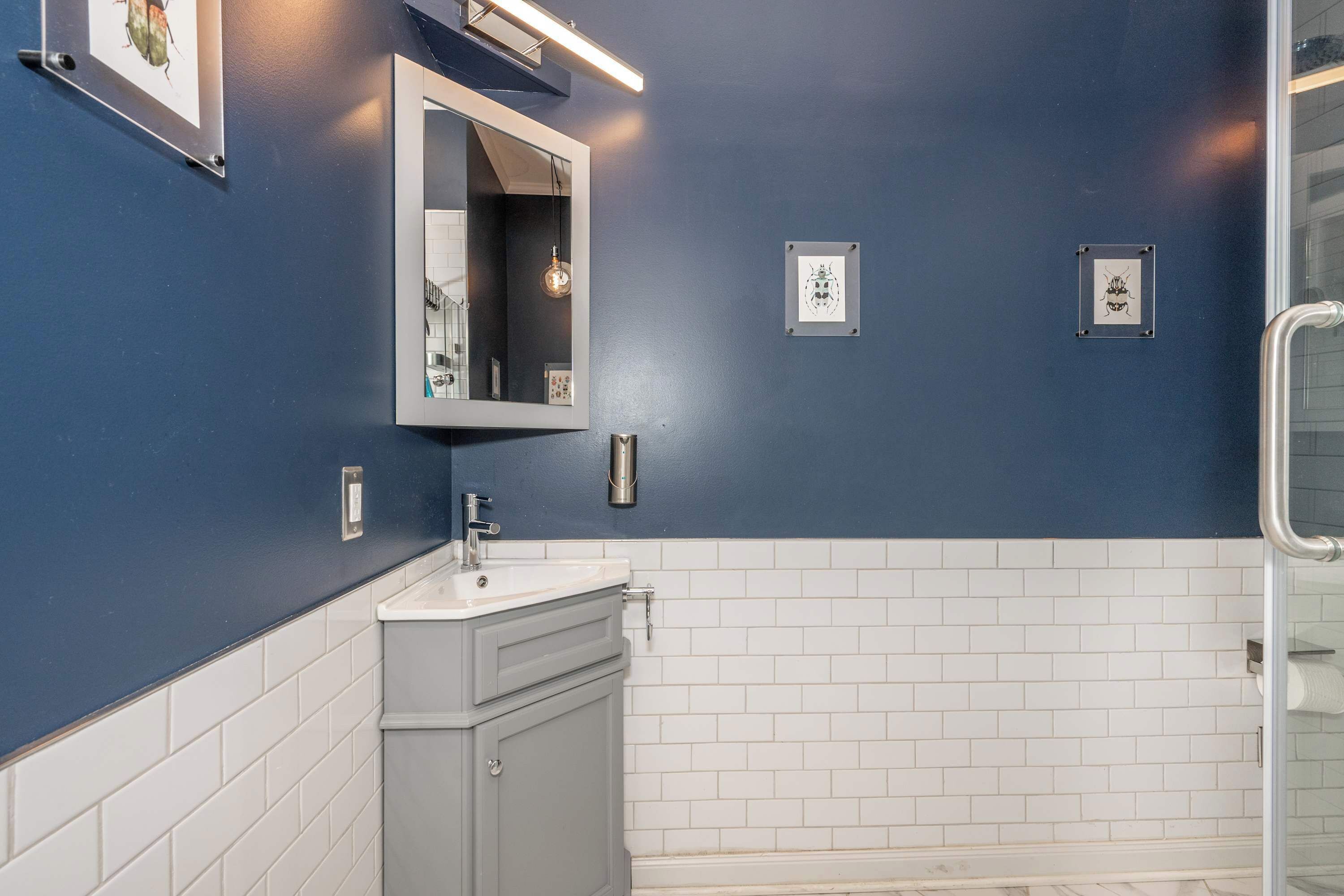 Bagno con parete blu, pavimento piastrellato bianco, lavabo, specchio e quadri appesi al muro.