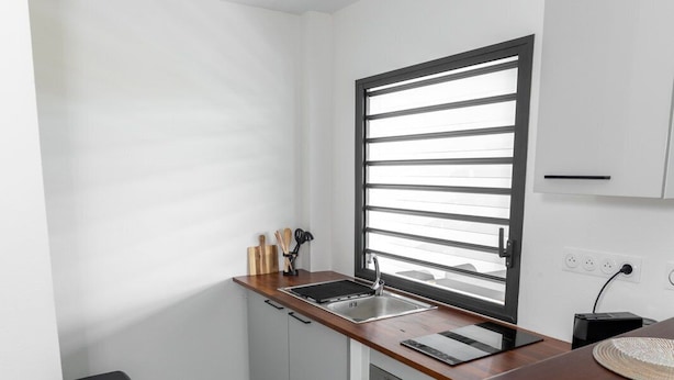 A kitchen with a wooden countertop, a window with horizontal blinds, a sink, and a trash bin. - Le Nid de Thalys  Appartement de charme (Cayenne)