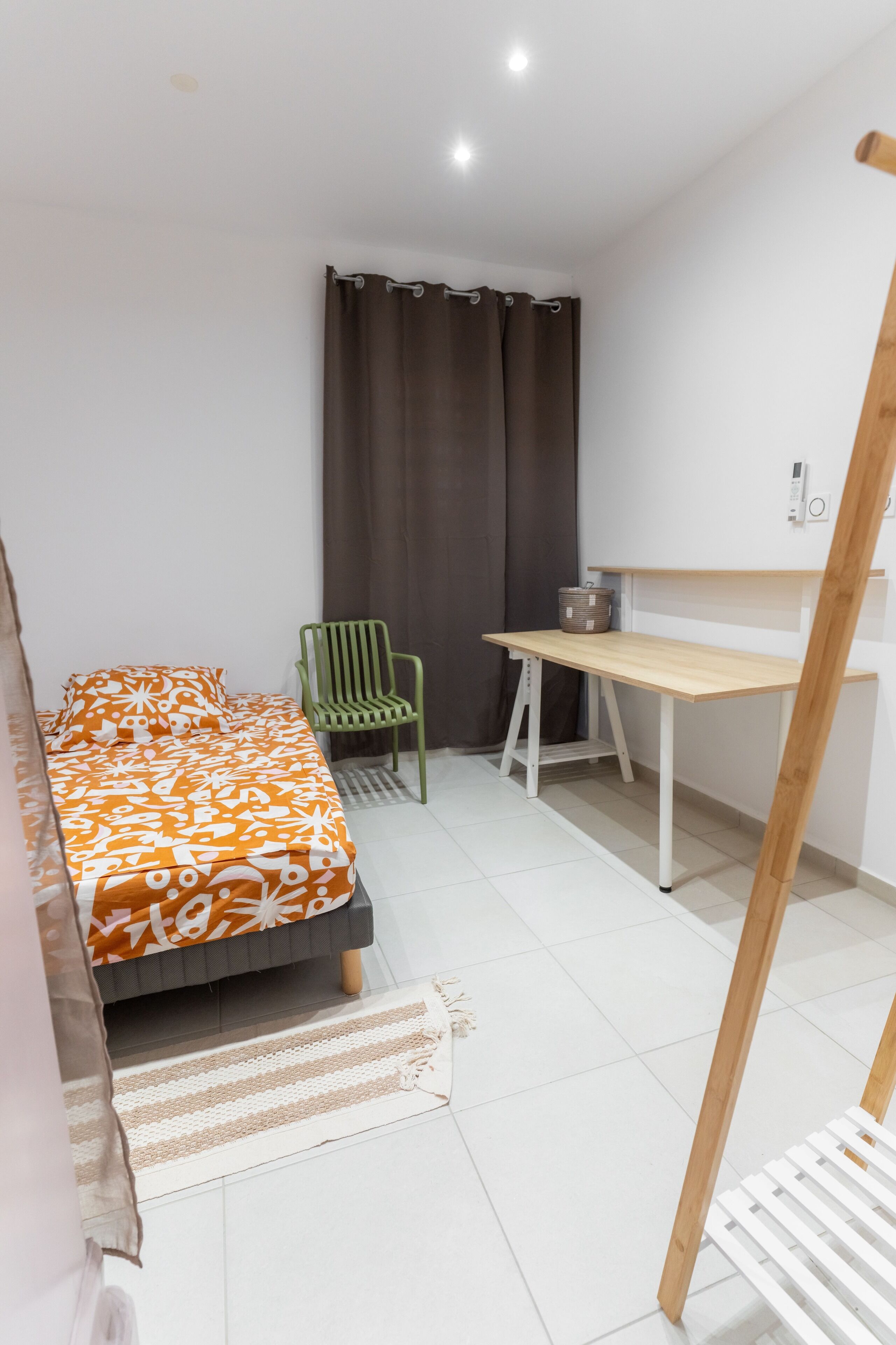 Een kleine, goed verlichte kamer met een bed, een bureau, een stoel en een raam met gordijnen.