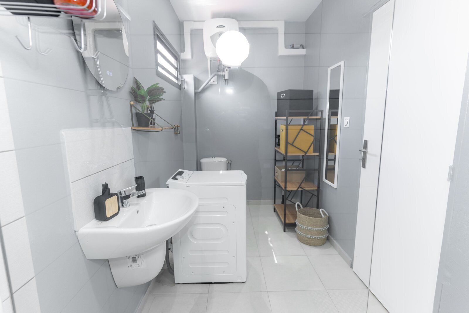 A modern bathroom with a white sink, toilet, and shower area. There is a mirror above the sink, a shelf with toiletries, and a hanging light fixture.