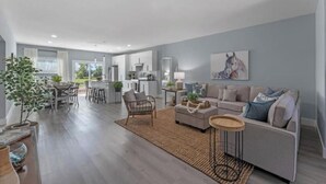 A spacious living area with a sofa, coffee table, and a dining table set. There is a kitchen in the background with modern appliances.
