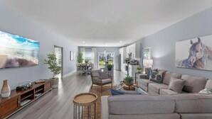 A spacious living area with a sofa, a dining table, and chairs. There is a TV mounted on the wall and a painting above the sofa.
