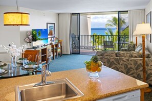A hotel room with a kitchen area, dining setup, and a balcony with a view of the ocean.