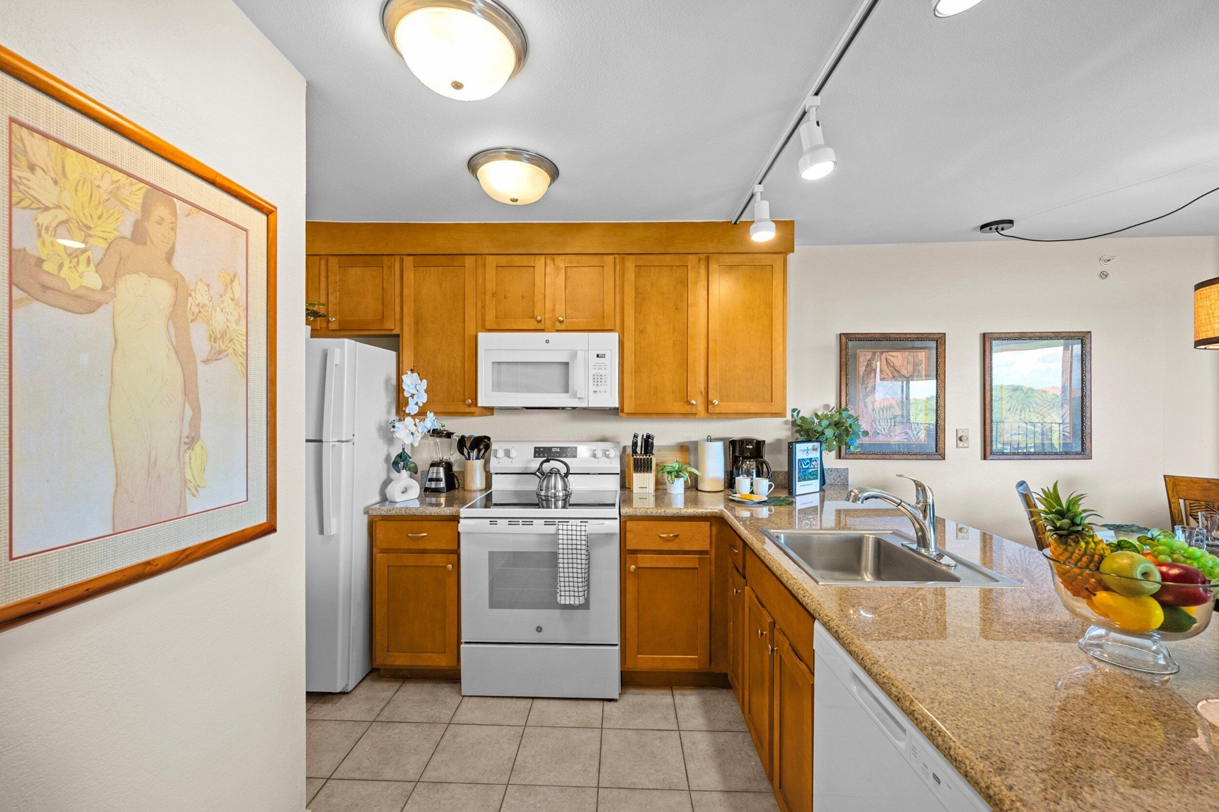A modern kitchen with wooden cabinets, a white refrigerator, a microwave, an oven, and a sink. There is a fruit bowl on the counter and a framed picture on the wall.