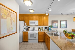 A modern kitchen with wooden cabinets, a white refrigerator, a microwave, an oven, and a sink. There is a fruit bowl on the counter and a framed picture on the wall.