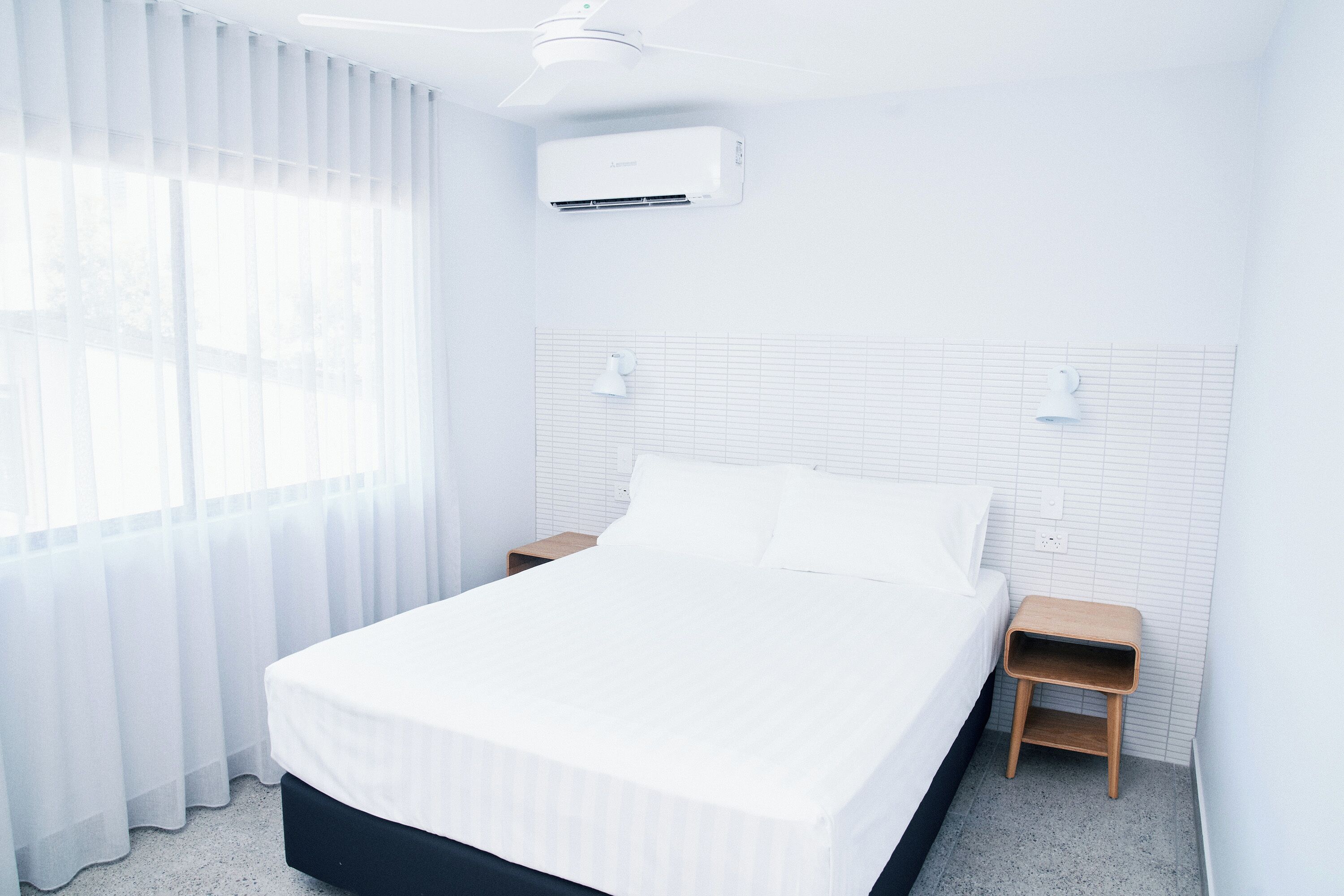 A hotel room with a bed, bedside tables, an air conditioning unit, and a window with curtains.