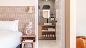 A modern hotel room with a bed, bedside table, and a bathroom with a sink and mirror.