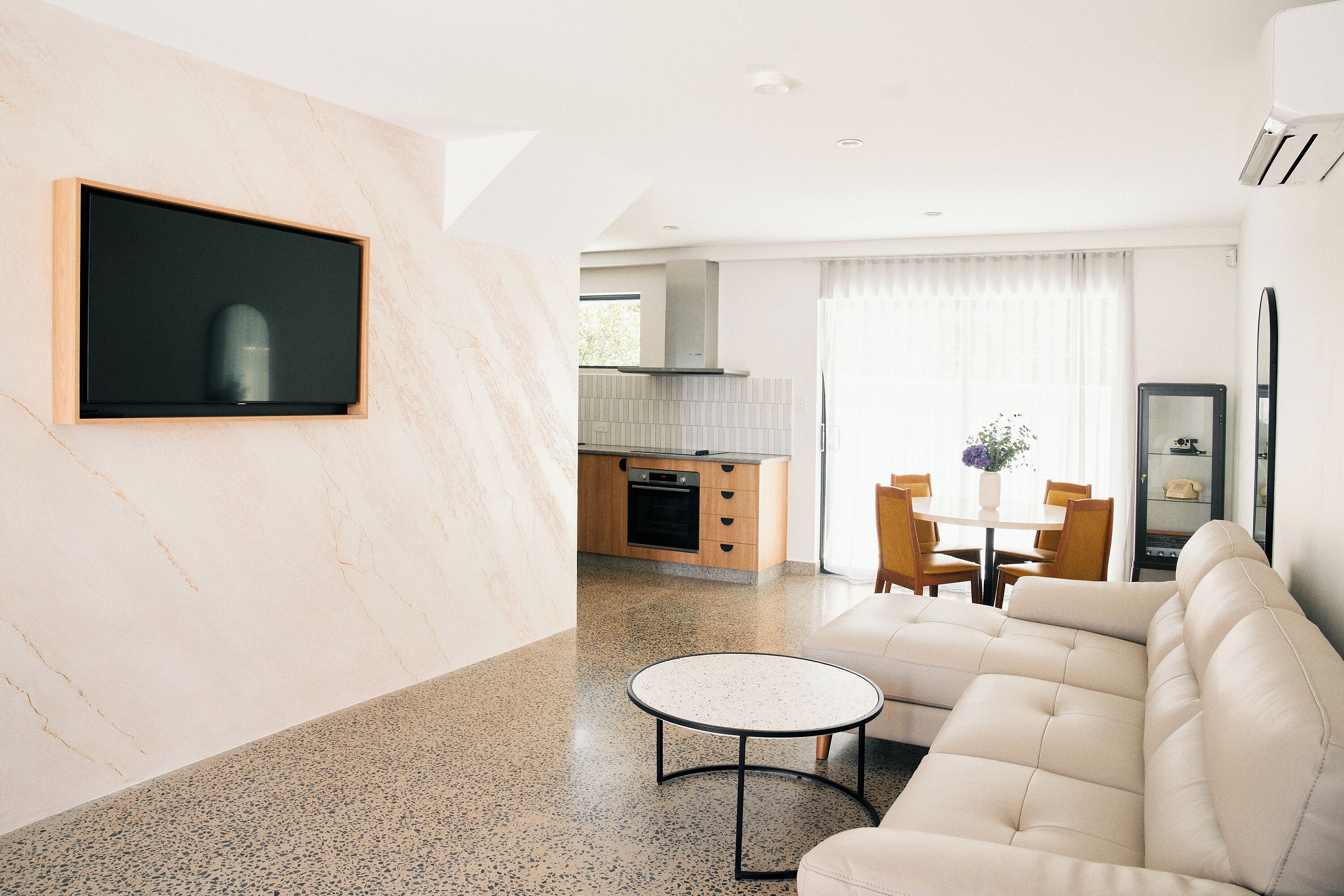 A modern living room with a sofa, a round coffee table, a TV mounted on a marble wall, and a kitchen in the background.