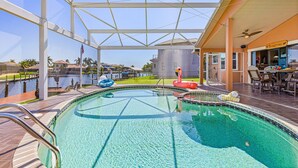 A swimming pool with a glass ceiling, a flamingo float, and a view of a lake and buildings in the background. - Sail into Paradise with Pool, Spa & Dockside Living - Captain Jack's Landing (Cape Coral)