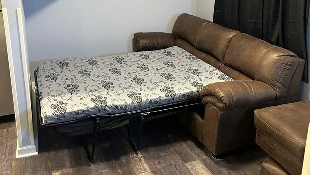 A compact room with a brown leather sofa, a small table with a patterned tablecloth, a mounted TV, and a ceiling-mounted air conditioner. - The Rustic Railroad Studio     5 Min to HWY 63            Kitchen + Washer/Dryer (Renick)