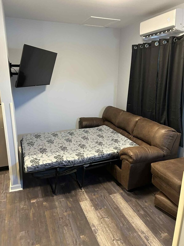 Ein gemütlicher Raum mit einer braunen Ledersofa, einem kleinen Tisch mit gemustertem Tischläufer, einem an der Wand montierten Fernseher und einer Decken-Klimaanlage.