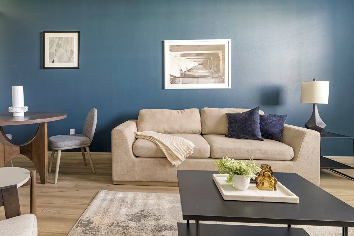 A living room with a blue accent wall, a beige sofa, a round wooden table, a grey armchair, and a black coffee table with decorative items.