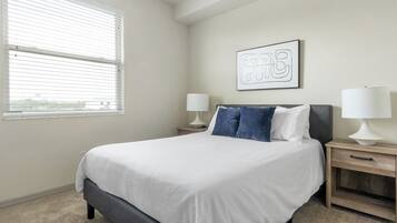 A bedroom with a bed, two bedside tables with lamps, a window with blinds, and a wall-mounted artwork.