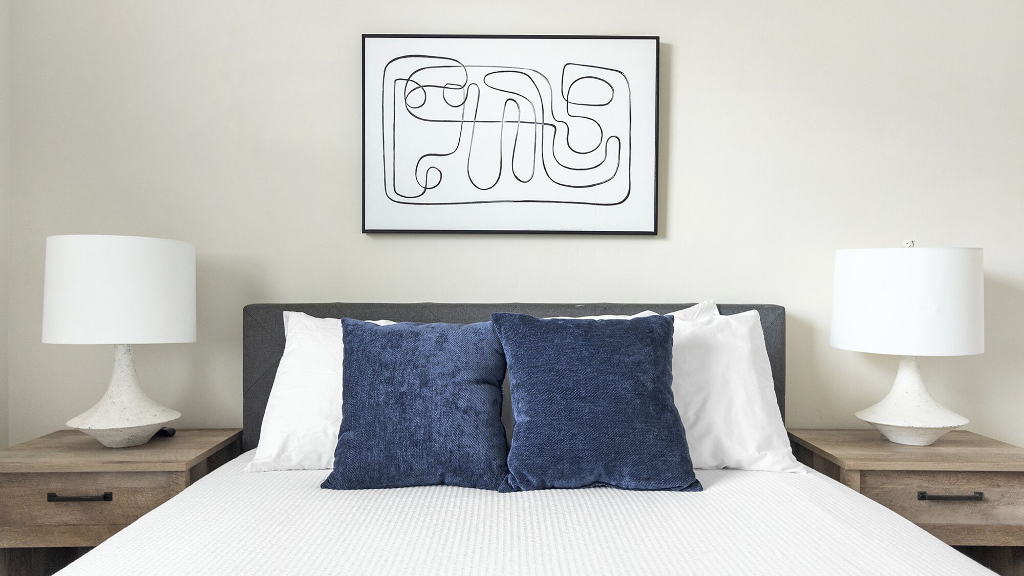 Una cama bien hecha con almohadas blancas y azules, dos lámparas de noche y una obra enmarcada en la pared.