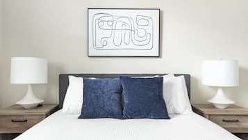 Una cama bien hecha con almohadas blancas y azules, dos lámparas de noche y una obra enmarcada en la pared.