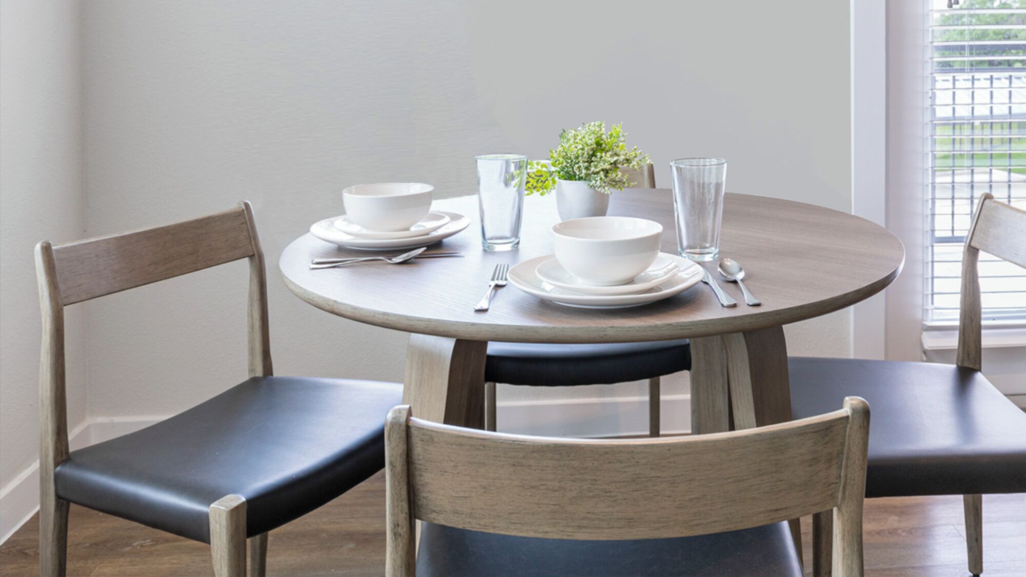 A round dining table with four chairs, white plates, bowls, glasses, and cutlery.