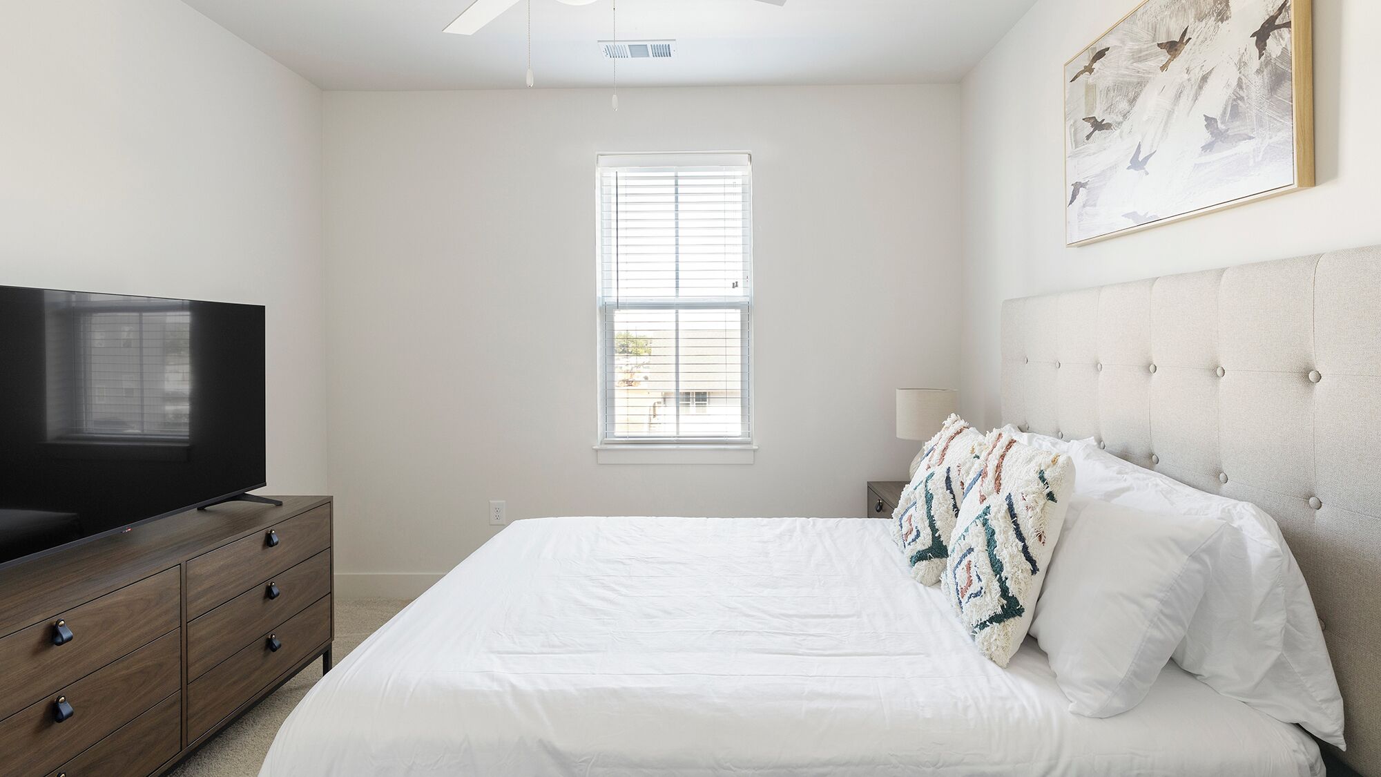 A bedroom with a bed, a TV on a dresser, a window, and a painting on the wall.