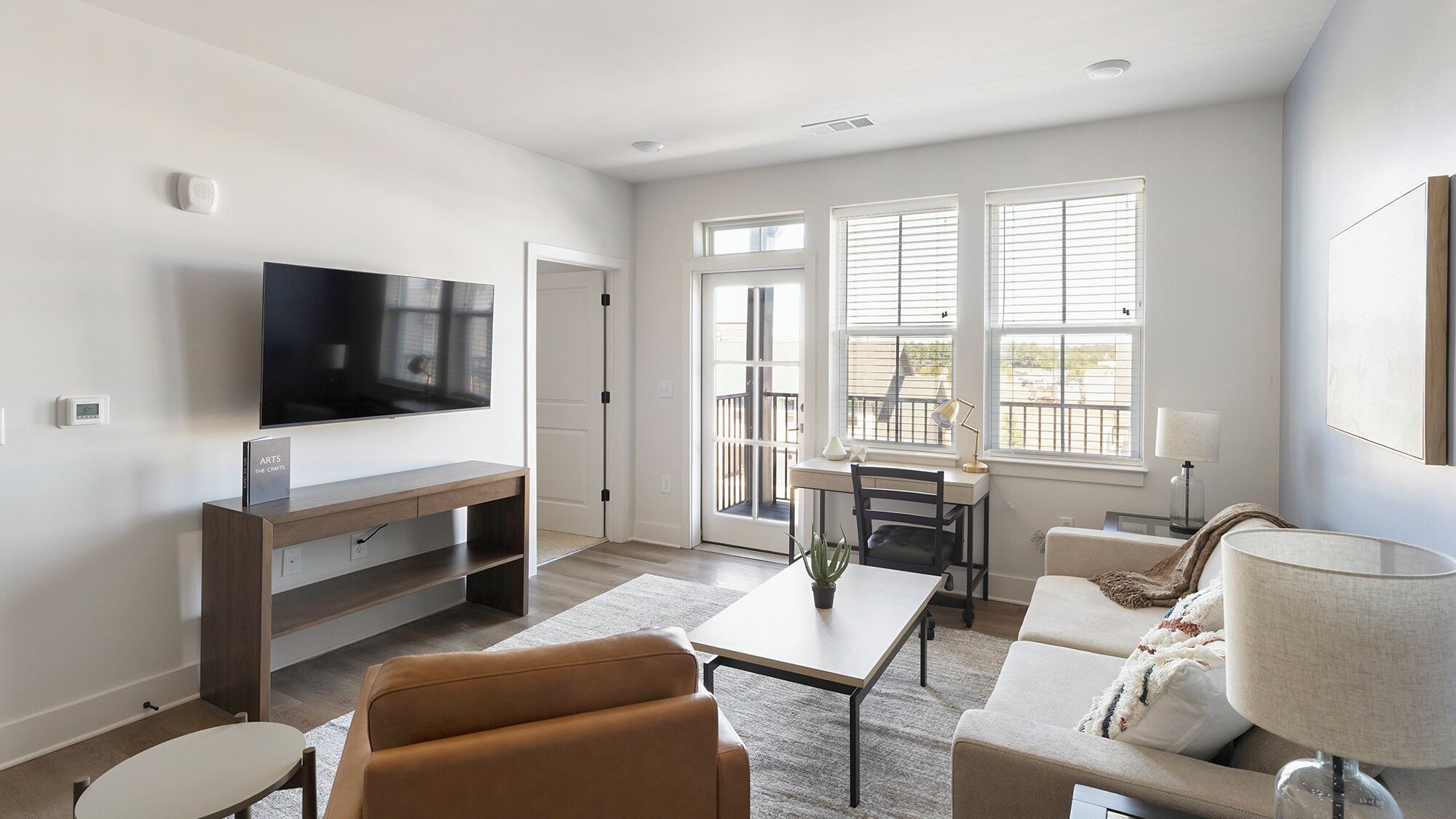 A modern living room with a flat-screen TV, a dining area with a table and chairs, and large windows providing natural light.
