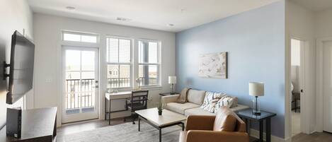 Una sala moderna con un sofá, sillones, una mesa de centro y un televisor. Se ve un balcón a través de las ventanas.