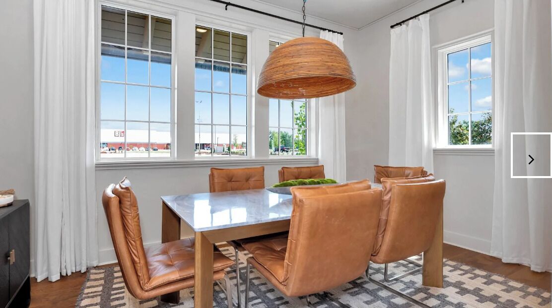 A dining area with a large table, six chairs, a hanging light fixture, and large windows with white curtains.