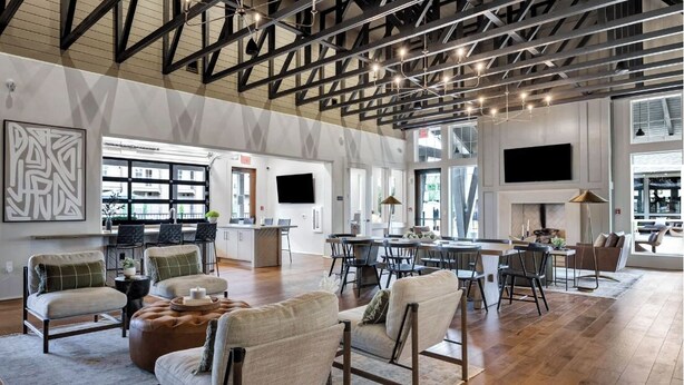 A spacious living area with high ceilings, exposed beams, and a large TV mounted on the wall. There are multiple seating areas with sofas and chairs, and a dining table with chairs. - Landing Apartments At Town Madison (Madison)