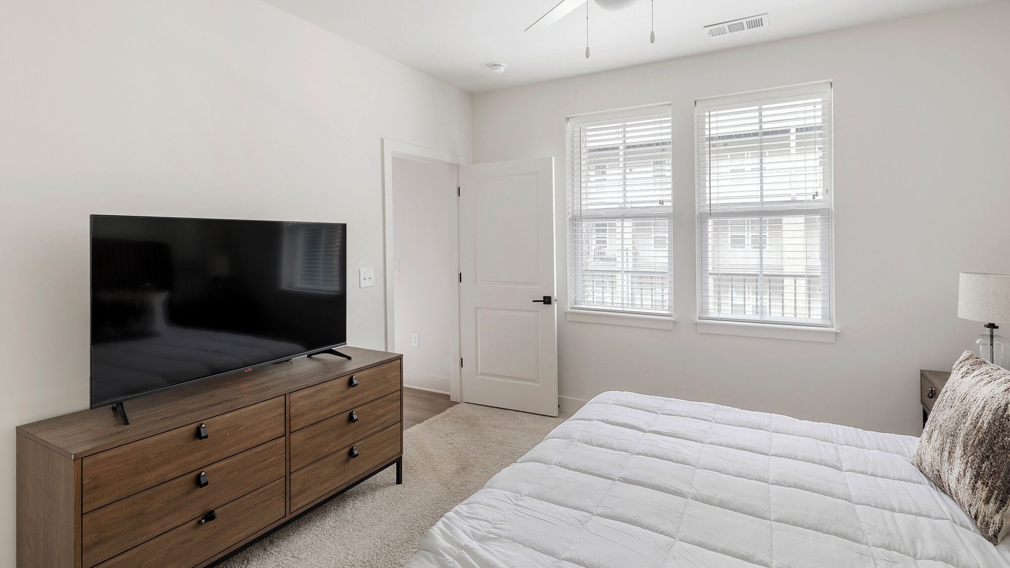 Un dormitorio con una cama, un aparador de madera con televisor, una puerta y ventanas con persianas.