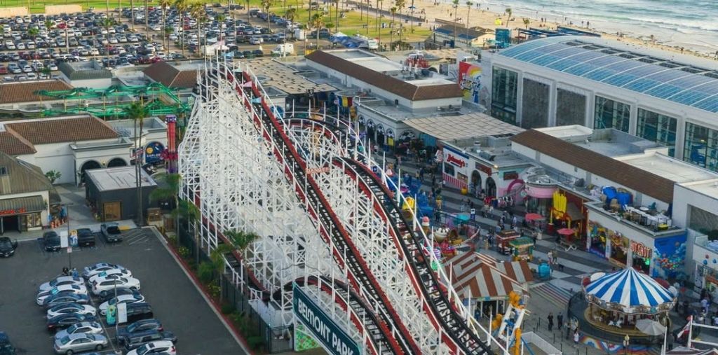 A view of Belmont Park with a roller coaster, shops, and a parking lot.
