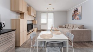 A modern kitchen with wooden cabinets, a dining table with chairs, a sofa, and a painting on the wall. - Osiedle Piastów Charming Apartment (Kraków)