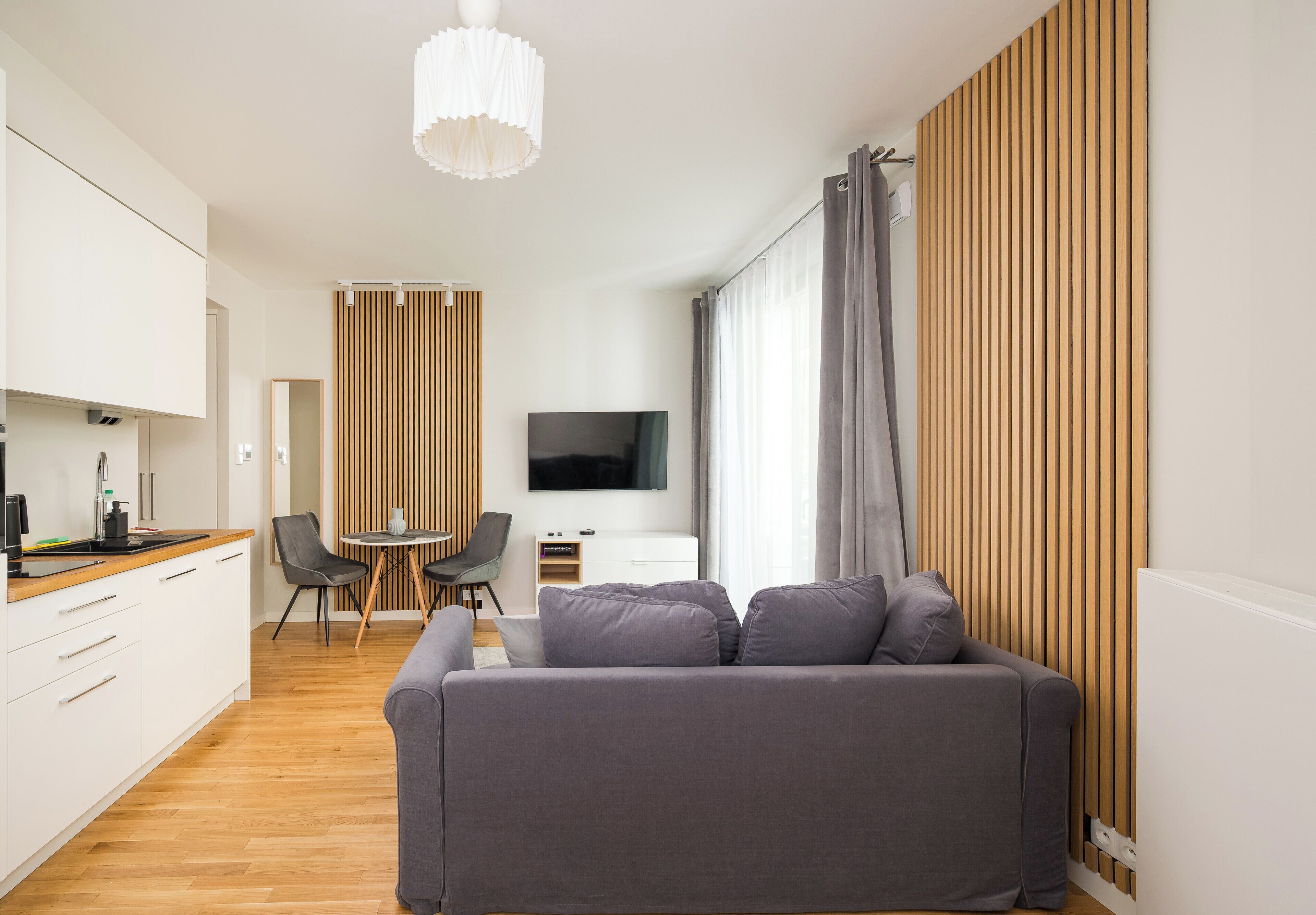 Un salon moderne avec un canapé gris, un téléviseur à écran plat, un coin repas comprenant une table et des chaises, et une cuisine dotée de meubles blancs et d’un plan de travail en bois.