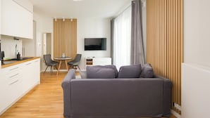 A modern living room with a grey sofa, a flat-screen TV, a dining area with a table and chairs, and a kitchen with white cabinets and a wooden countertop. - Krasiński Garden Deluxe Studio (Warszawa)