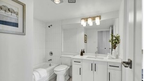 Un baño moderno con lavamanos, inodoro y bañera blancos. Hay un cuadro en la pared y un espejo sobre el lavamanos.