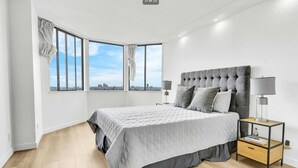 Un dormitorio moderno con una cama grande, dos mesitas de noche y vistas a la ciudad a través de amplios ventanales.