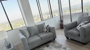 Un salón moderno con un sofá gris, un tapete estampado y vistas a un paisaje urbano a través de grandes ventanales.