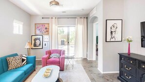 Ein Wohnzimmer mit einem blauen Sofa, einem pinken Sessel, einem weißen Couchtisch und einer schwarzen Kommode. An den Wänden befinden sich Bilder in Rahmen und es gibt einen Deckenventilator.