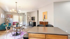 Ein modernes Wohnzimmer mit Essbereich, Kücheninsel und Fernseher. An den Wänden befinden sich Bilder in Rahmen, und es gibt einen Deckenventilator.