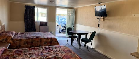 Habitación de hotel con dos camas, una pequeña mesa con sillas, un televisor montado en la pared y vista a coches estacionados afuera.
